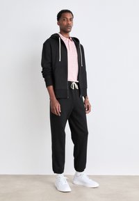 Sudadera negra con cremallera y cordones, combinada con una camiseta polo rosa claro y pantalones jogger negros, complementados con zapatillas blancas. Texturas suaves en toda la indumentaria.