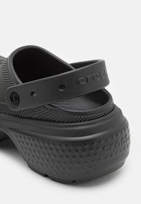 Sabot Crocs noir avec semelle texturée et bride de talon réglable embossée du nom de la marque sur fond blanc.