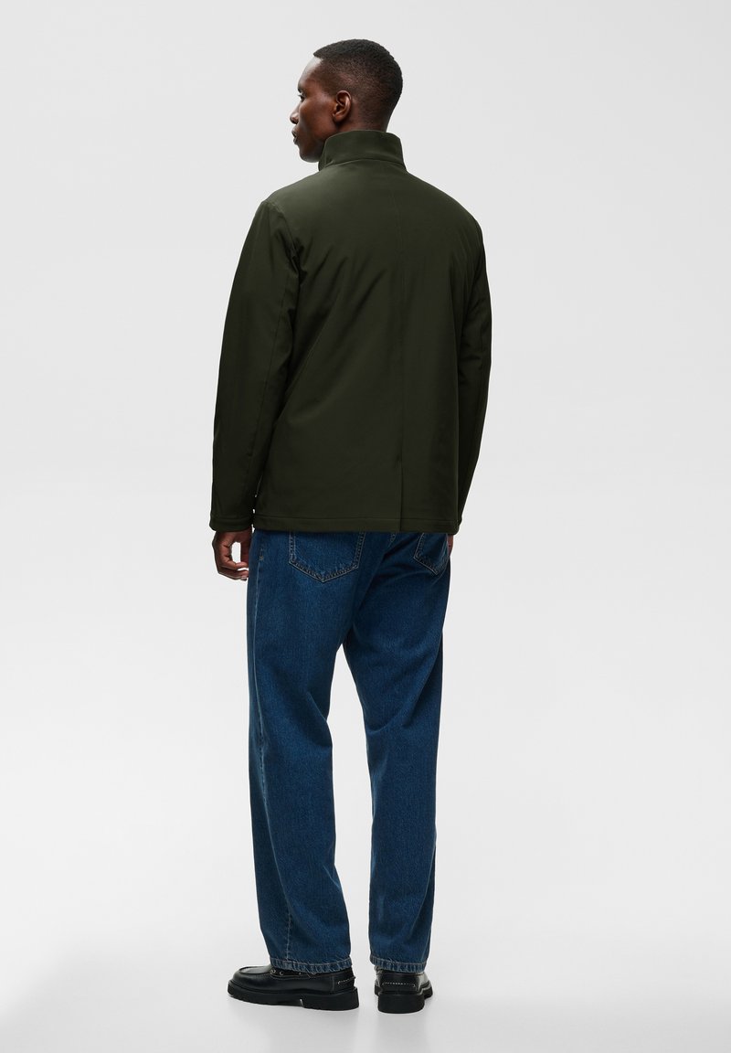 Homme debout, de dos, portant une veste vert foncé avec le col relevé, un jean bleu et des chaussures noires sur un fond clair uni.