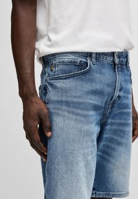 Pantalones cortos de mezclilla azul claro con un lavado desteñido, que cuentan con un diseño clásico de cinco bolsillos y un cierre de botón. Tejido texturizado con sutiles detalles de costura.