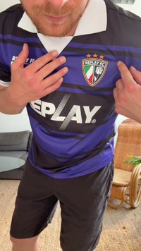 Futbolo marškiniai su tamsiai mėlynomis ir purpurinėmis juostomis, balta apykaklė, su "REPLAY" logotipu ir "REPLAY FC" ženklu, kuriame yra trys žvaigždutės.