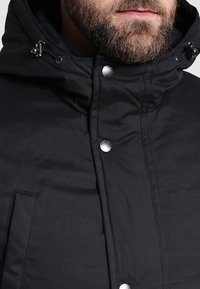 Homme barbu portant une veste d'hiver noire à capuche avec des boutons-pression et des cordons ajustables près du col.