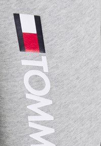 Tela de algodón gris con un logotipo textil brillante. Presenta un texto vertical en blanco "TOMMY" y un logotipo rectangular en rojo, blanco y negro en el lateral.