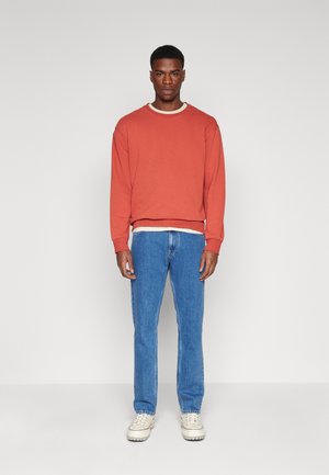 Sudadera naranja de manga larga con cuello redondo, superpuesta sobre una camisa blanca, combinada con jeans azules y zapatillas de color crema.