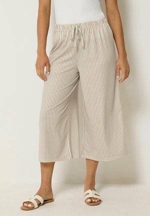 Kvinde iført beige og hvidt lodretstribet, vidbenet, cropped bukser med snøre i taljen og hvide slippers.