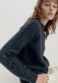 Marineblauer Strickpullover mit geripptem Ausschnitt und strukturiertem Zopfmuster an den Ärmeln. Weiches Material mit lockerer Passform.