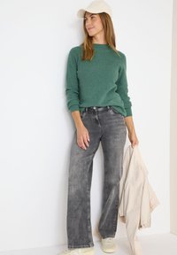 Pull en maille côtelée vert, jean large gris, et une veste beige clair tenue d'une main, accompagnés d'une casquette blanche et de baskets.