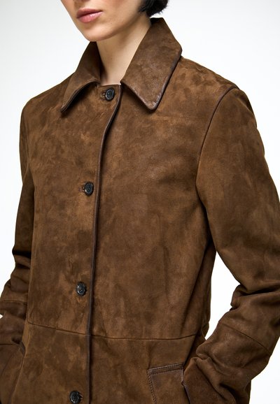 Femme portant un manteau boutonné en daim marron avec col, les mains dans les poches.