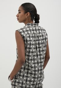 Donna con un chignon intrecciato, che indossa una canotta con un motivo geometrico bianco e nero, guarda a sinistra con una mano nella tasca su uno sfondo neutro.
