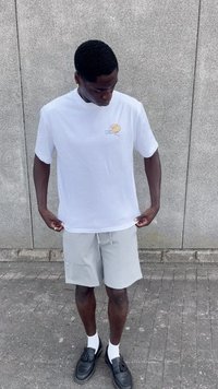 Camisa de algodón blanca con un pequeño gráfico amarillo en el lado izquierdo del pecho, combinada con pantalones cortos gris claro y zapatos negros sin cordones.