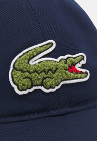 Gorra azul marino con un logo de cocodrilo bordado en verde con acentos rojos, contorneado en blanco. Confeccionada en tejido texturizado.