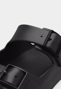 Close-up van een zwarte sandaal met twee verstelbare bandjes en een matte afwerking, met zichtbare gespen en gestructureerde oppervlakdetails.