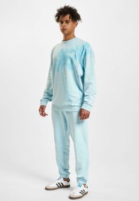 Light blue sweatshirt with palm tree design paired with matching joggers. Features ribbed cuffs and waistband, worn with white and brown sneakers.