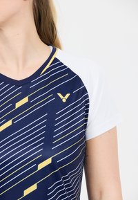 Nainen, jolla on päällään tummansininen urheilupaita, jossa on valkoisia ja keltaisia vinoja raitoja ja valkoiset hihat, näkyy kaulasta ylöspäin kyynärpäähän.