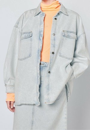 Osoba nosząca jasnoniebieską, oversize dżinsową kurtkę i spódnicę z brzoskwiniowym golfem, stoi z jedną ręką w kieszeni kurtki.