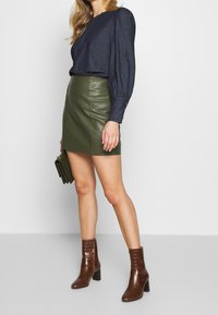 Top con maniche a sbuffo color navy, minigonna in pelle verde oliva e stivaletti alla caviglia in pelle effetto coccodrillo marroni; borsa a tracolla in mano.