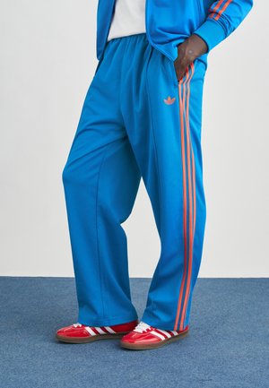 Pantalones deportivos azules con rayas laterales naranjas, cintura elástica y corte holgado. Combinados con zapatos rojos con rayas blancas.