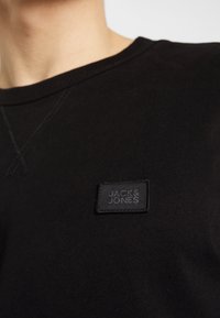 Černá mikina s kulatým výstřihem vyrobená z měkké látky, s texturovanou černou nášivkou "JACK & JONES" vyšitou na prsou.