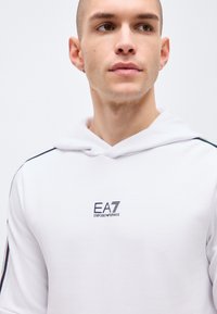 Joven con sudadera con capucha blanca Emporio Armani EA7 con logo negro, mirando hacia la derecha, fondo neutro.