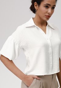 White, textured short-sleeve blouse with a collared neckline, front button closure, and a relaxed fit. Worn with high-waisted beige pants.