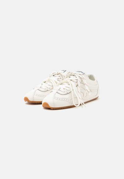 Paire de baskets basses blanches avec semelles beige et lacets blancs noués lâchement sur un fond gris clair uni.