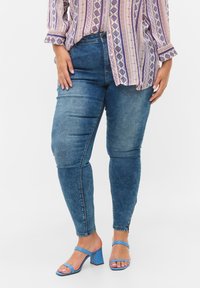 Jeans ajustados de mezclilla azul medio, con una textura suave y un detalle de cremallera en los tobillos. Combinados con una blusa estampada y sandalias azul con tacón cuadrado.
