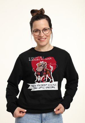 Kvinde med briller og hår samlet i en knold smiler, mens hun bærer en sort sweatshirt med en tegneseriehund og tekst om ansvarsfuldt kæledyrsejerskab.