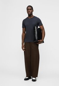 Homme portant un t-shirt noir, un pantalon large marron, des chaussures noir et blanc, tenant un grand sac fourre-tout noir sur un fond uni.