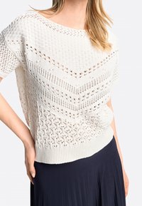 Hvid strikket top med hulmønstre, løstsiddende ærmer og ribbet kant. Båret over en marineblå plisseret nederdel.