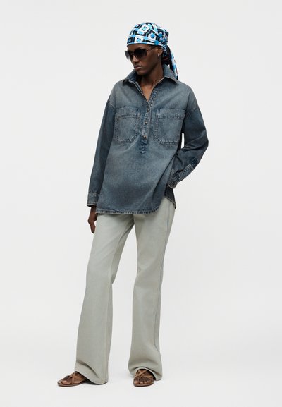 Άτομο με σκούρο πουλόβερ από τζιν, ανοιχτόχρωμο τζιν με φαρδιά πόδια, μπλε μαντίλι με μοτίβα στο κεφάλι, γυαλιά ηλίου και καφέ σανδάλια, στέκεται με το ένα χέρι στην τσέπη.