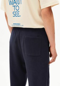 Marineblauwe joggingbroek met een gestructureerde afwerking, voorzien van een open achterzak en een elastische tailleband. De outfit wordt aangevuld met een ivoorkleurig t-shirt.