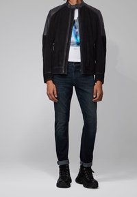 Svart mockabomberjacka med läderdetaljer, ihop med mörka jeans och svarta snörstövlar. Jackan har hög krage och dragkedjedetaljer.