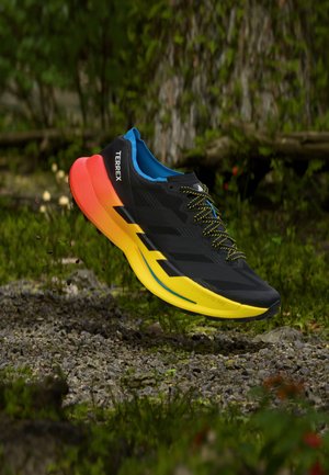 Zapato deportivo negro con parte superior texturizada, suela amarilla y naranja, y detalles azules, con cordones negros y un logo "TERREX" en el lateral.