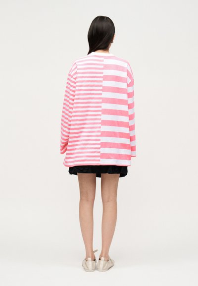 Γυναίκα στέκεται με την πλάτη προς τον θεατή, φοράει υπερμεγέθη ροζ και λευκή ριγέ μπλούζα, μαύρη φούστα και λευκά αθλητικά σε μονόχρωμο φόντο.