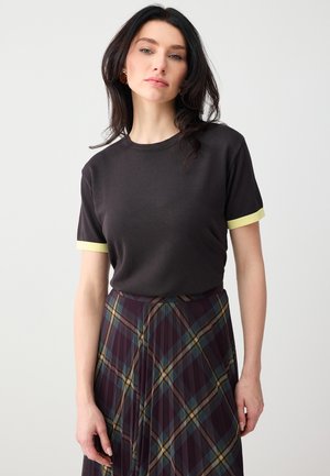 Donna con capelli scuri che indossa una maglietta nera con bordi delle maniche gialli e una gonna plissettata a quadri scuri, in piedi davanti a uno sfondo semplice.