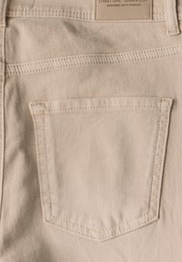 Beige denim kangas, jossa näkyvä takatasku, suorassa muodossa, hienovarainen rakenne ja valkoiset ompelu yksityiskohdat. Tulostettu merkki ylhäällä.