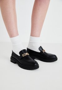 Melni ādas loafers ar masīvu zoli un zelta detaļām. Valkāti ar baltām kokvilnas zeķēm, akcentējot gludo tekstūru un eleganto dizainu.