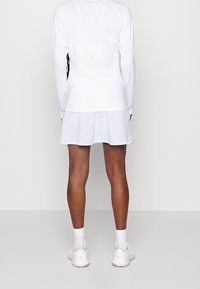 Top blanco de manga larga combinado con una falda acampanada de un gris claro. El top tiene una textura suave; la falda presenta un patrón sutil. Se ven deportivas blancas.