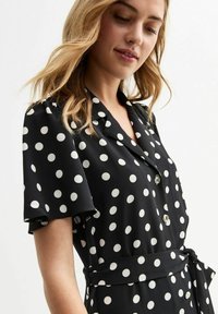 Vestido negro con lunares blancos. Presenta mangas cortas, un cuello, botones frontales y una cintura atada para un ajuste ceñido.