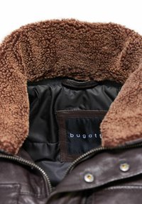 Bruine leren jas met een zachte bruine schapenwol kraag en een zwarte binnenvoering. Voorzien van een logo patch en een metalen ritsluiting.
