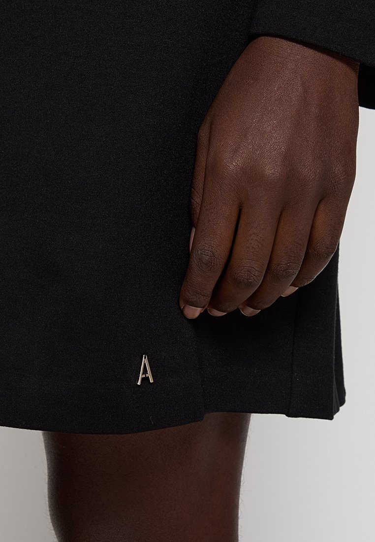 Czarna odzież o teksturowanym wykończeniu, z widoczną dłonią trzymającą rąbek. Akcent metalowej litery "A" w pobliżu rąbka.
