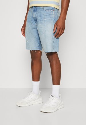Shorts en denim bleu clair avec un ourlet effiloché, présentant un design usé. Associés à des baskets blanches et des chaussettes montantes.