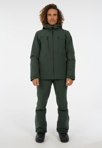 Groene skijas met hoge kraag, twee ritsborstzakken en bijpassende groene skibroek, gemaakt van weerbestendige stof. Zwarte laarzen.