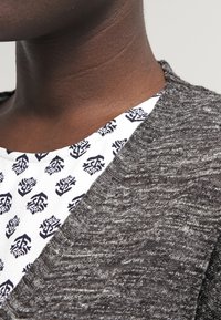 Suéter gris de punto con escote en V, sobre una camisa blanca con un patrón floral azul. Primer plano del escote y la textura.