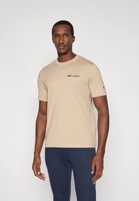 Μπεζ βαμβακερό t-shirt με κοντά μανίκια, στρογγυλή λαιμόκοψη και μικρό μαύρο λογότυπο Champion στο στήθος. Φοριέται με μπλε ναυτικό παντελόνι.