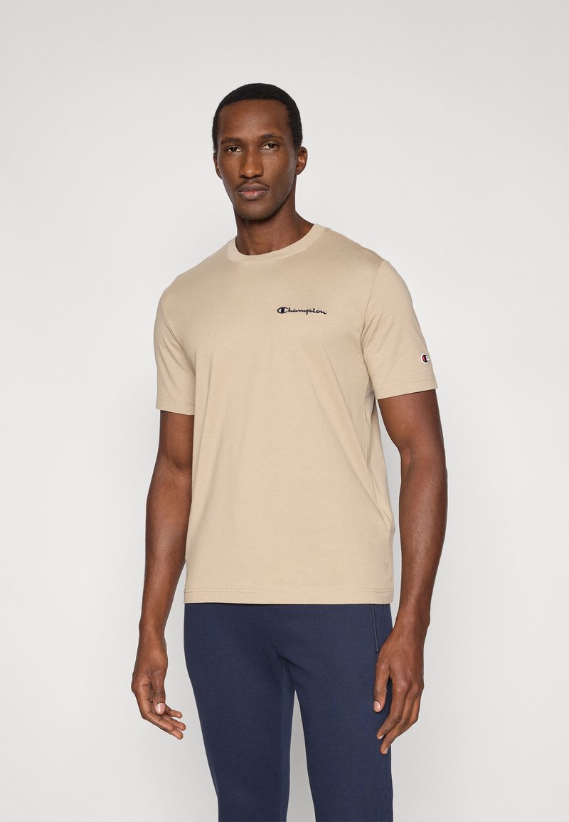 Μπεζ βαμβακερό t-shirt με κοντά μανίκια, στρογγυλή λαιμόκοψη και μικρό μαύρο λογότυπο Champion στο στήθος. Φοριέται με μπλε ναυτικό παντελόνι.