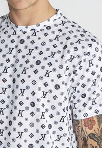 Camiseta blanca de manga corta con un patrón negro de varios logotipos, letras y símbolos, con cuello redondo y una textura suave.
