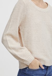 Helle beige gestrickte Sweater mit weitem Ausschnitt, langen Ärmeln und gerippten Bündchen. Texturiert mit einer lockeren Passform. Kombiniert mit blauen Jeans.