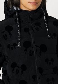 Chaqueta acolchada negra con un sutil patrón de Mickey Mouse en toda la prenda, cierre frontal con cremallera y cordones contrastantes con texto. Textura suave.