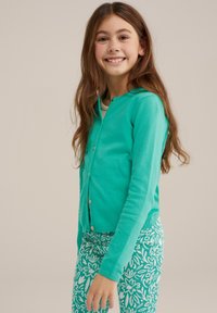 Tirkīza krāsas garas piedurknes kardigāns ar noapaļotu kakla izgriezumu un zelta pogām, saskaņots ar zaļām legingām, kurām ir baltas ziedu raksts.